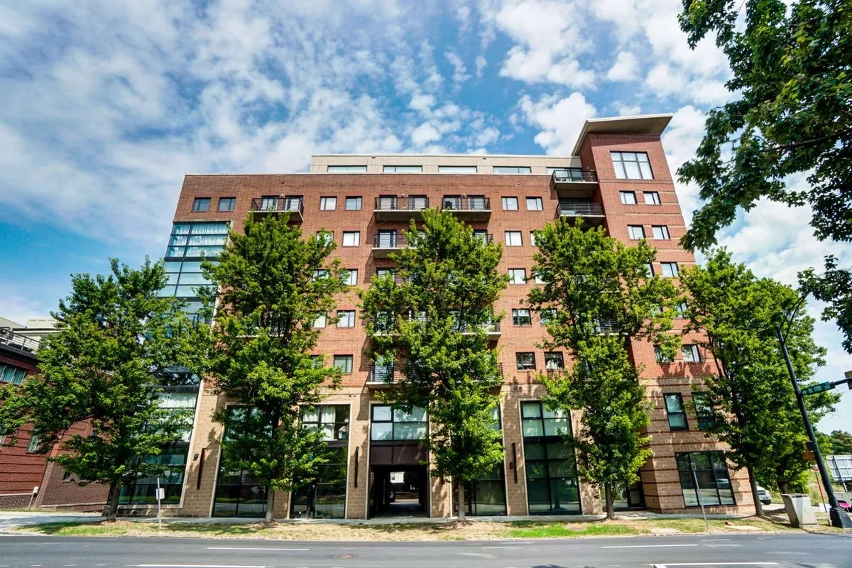 This exterior image highlights the modern brick architecture of the building, featuring expansive glass windows and a symmetrical line of trees leading toward the entrance. The visible balconies hint at private outdoor spaces, and the mood is welcoming, suggesting moments of relaxation and community gatherings.