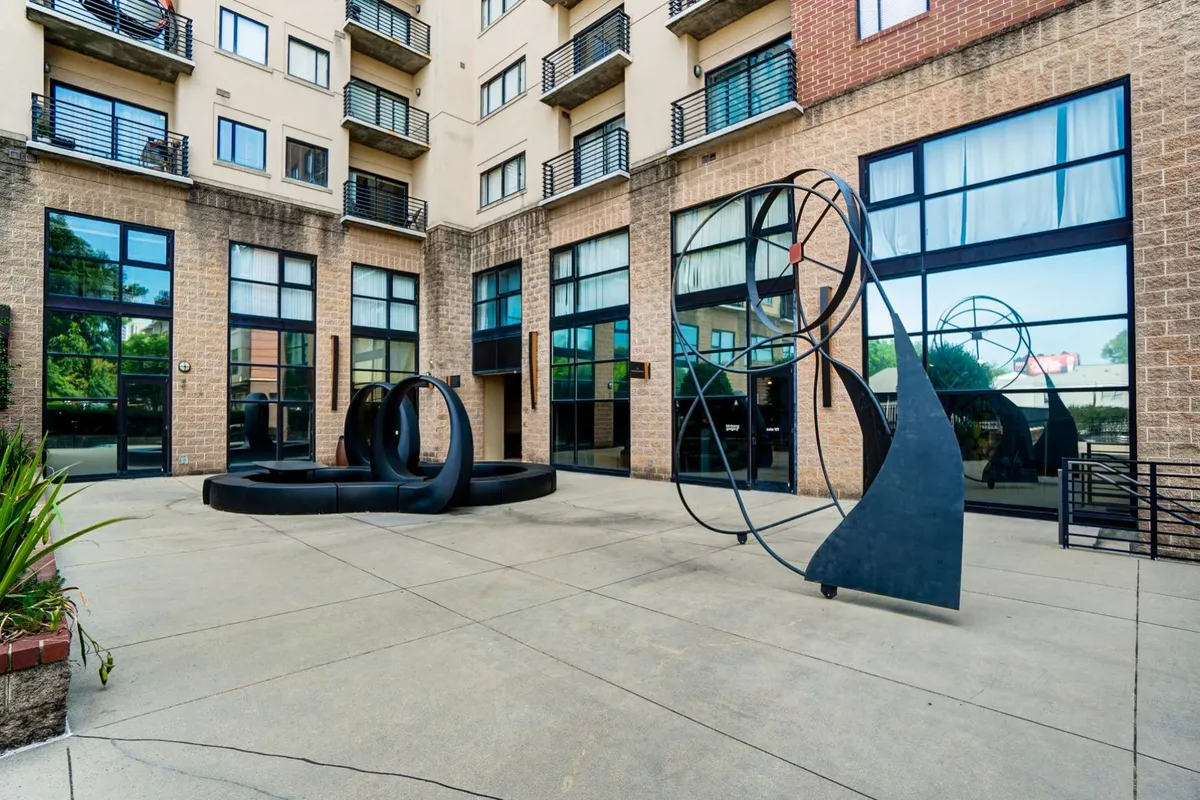 The image captures the building's exterior, focusing on artistic sculptures, large windows, and balconies. The communal courtyard area is highlighted, with a contemporary and inviting mood suggestive of social gatherings and relaxation during summer evenings.