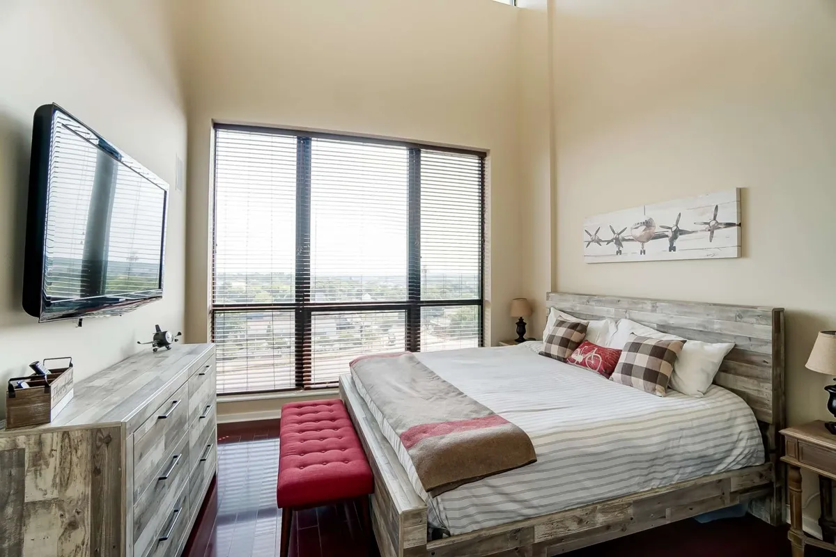 This image features a serene master bedroom with a large bed adorned with decorative pillows, a red ottoman at its foot, expansive windows with blinds, a wooden dresser, and a wall-mounted television. Sunlight streams through the windows, creating a tranquil, inviting atmosphere ideal for rest and relaxation.