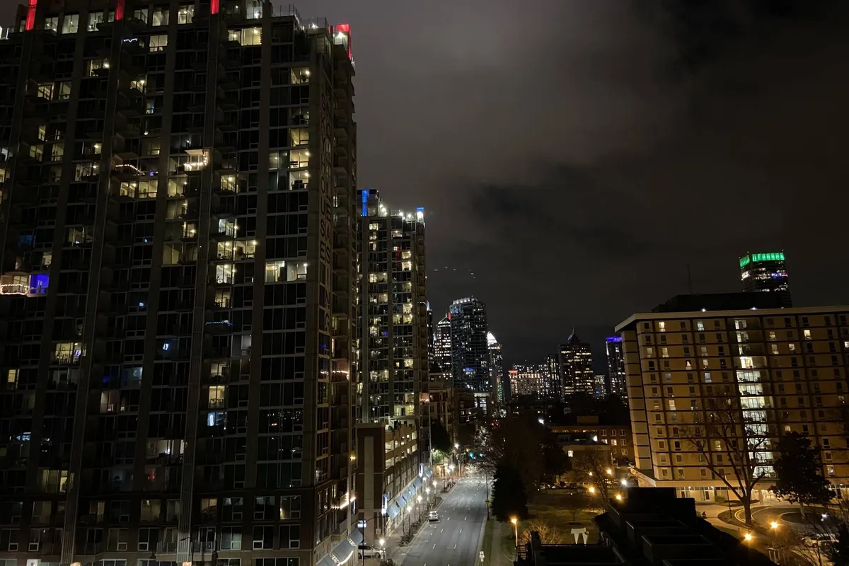 This exterior image features tall modern buildings illuminated against the night sky, with streetlights casting a vibrant glow. The city skyline is visible, and the lively street atmosphere hints at nightlife, evening events, and urban exploration.