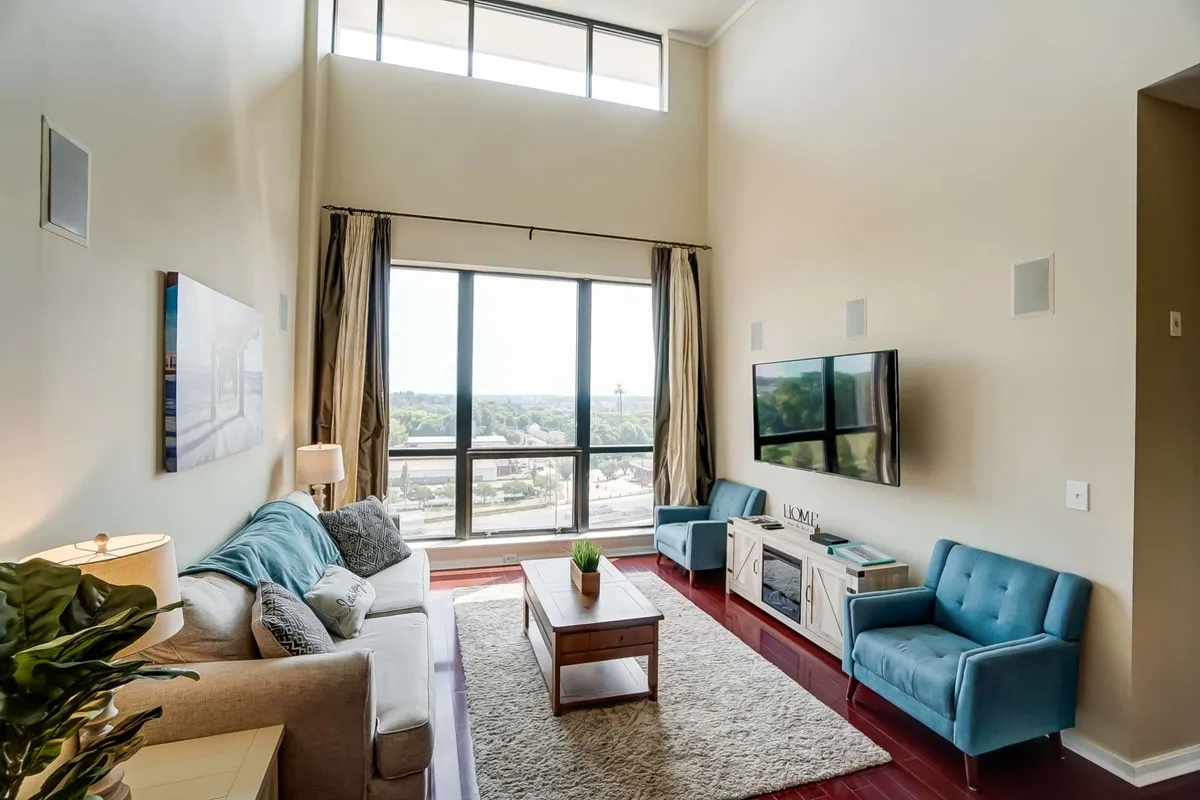 A spacious living room with large windows that flood the area with natural light, offering expansive views of the city skyline. The room features a comfortable sofa, a wall-mounted TV, stylish blue chairs, and a central coffee table, all arranged in an open, inviting layout. The decor is contemporary, with designer touches and a welcoming atmosphere ideal for relaxing or entertaining.