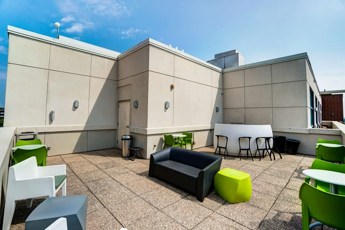 Pictured is a contemporary outdoor lounge area complete with a black sofa, green stools, white tables, and stylish lighting fixtures. The open-air setting invites relaxation, dining, and social interaction, while also offering the ambiance of sunset views and city lights.