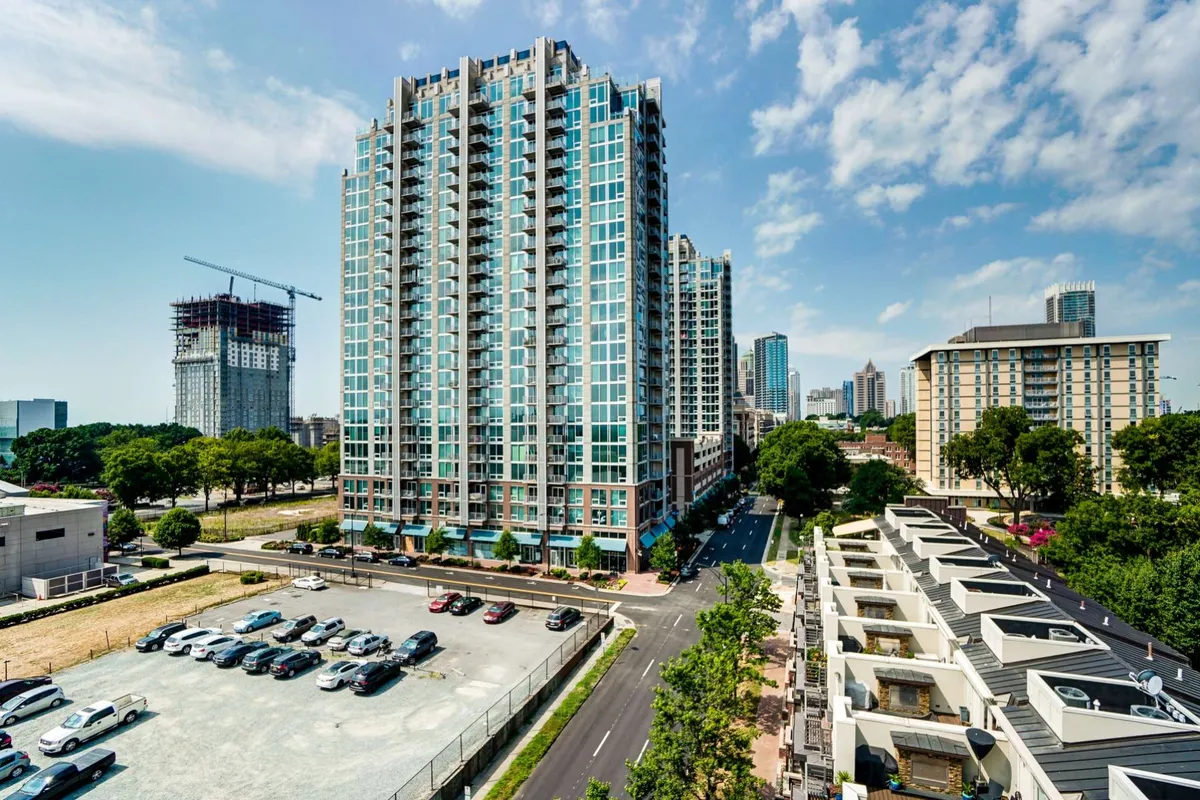 A dynamic exterior scene showing a tall residential building surrounded by city life, with green trees lining the street and a parking lot in view. The image conveys urban living with easy access to amenities and a sense of community.
