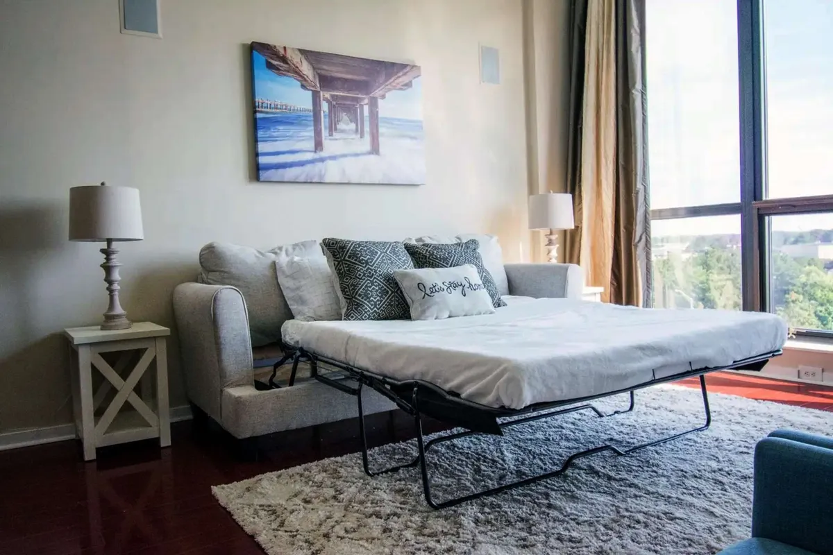 This cozy interior scene highlights a convertible sofa bed adorned with decorative pillows, stylish wall art, and warm lighting from table lamps. The space feels intimate, flexible, and perfect for reading or relaxing—ideal for accommodating guests or couples with different sleeping needs.